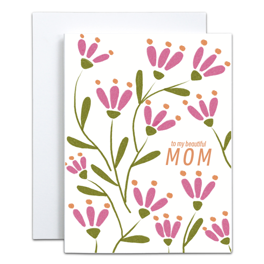A greeting card with a white background, decorated with pink flowers, green stems, and orange dots, with the text 'to my beautiful MOM' printed on the front. A white envelope is partially visible behind the card.