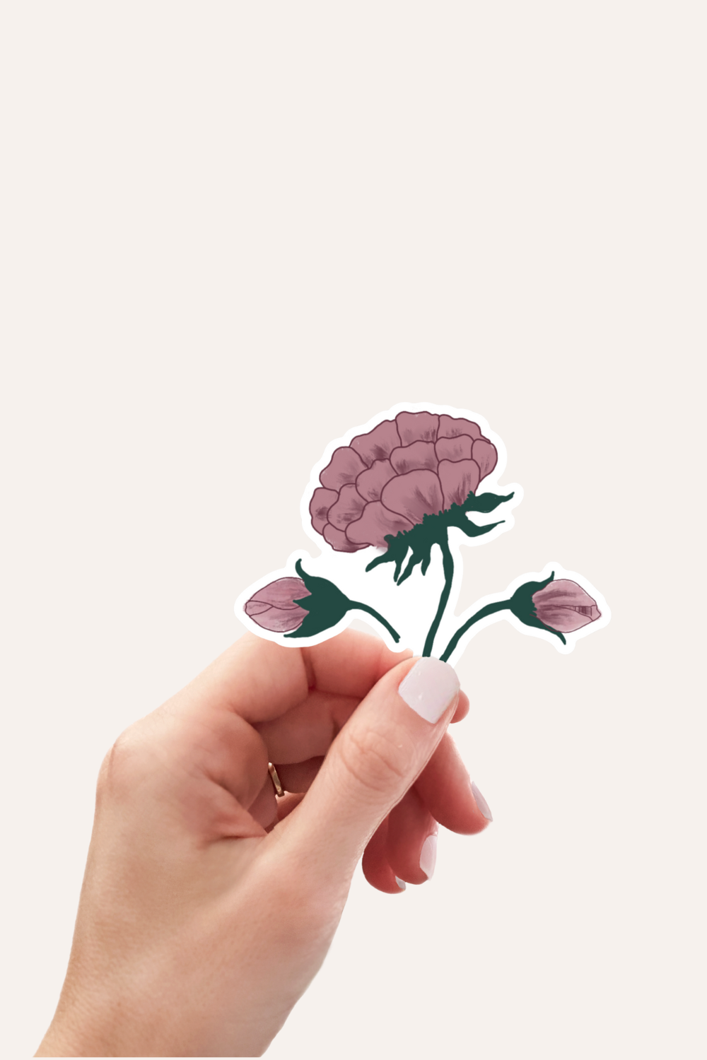 A hand with pale pink nail polish holds a die-cut sticker of a mauve-pink flower with green stems and buds against a solid off-white background.