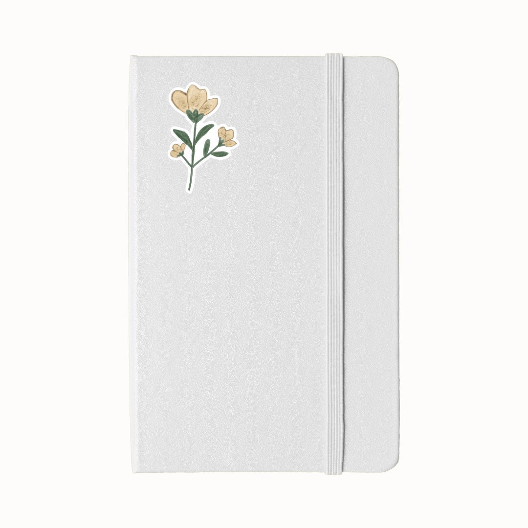 A white hardcover notebook with an elastic closure band and a sticker of a floral illustration on the upper left, shown against a plain white background.
